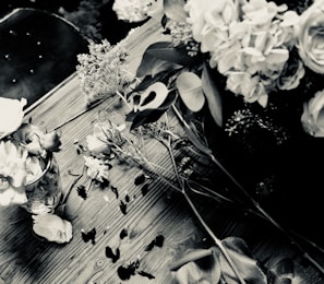 A black and white photograph of a wooden table with scattered flowers and petals. A vase holds some of the flowers upright. The composition includes leaves and a variety of floral elements, creating a rustic and natural setting.