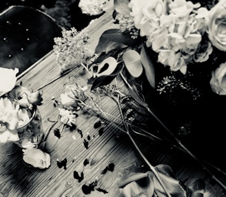 A black and white photograph of a wooden table with scattered flowers and petals. A vase holds some of the flowers upright. The composition includes leaves and a variety of floral elements, creating a rustic and natural setting.