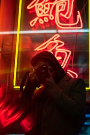 A person is taking a photo with a camera in front of brightly lit neon signs. The red and yellow neon signs illuminate the background, casting a vibrant glow and creating a contrast with the silhouette of the person.