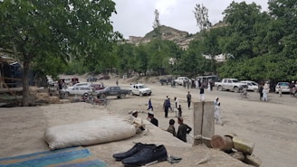 A dusty road in a rural area with several parked cars, including some off-road vehicles. Groups of people are gathered and conversing, and a few individuals are walking or cycling. Large trees and a hillside with scattered buildings are visible in the background. In the foreground, there are a pair of black shoes and a large sack, suggesting a casual setting.