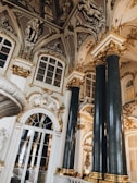 Ornate interior featuring large marble columns with gold accents and intricate moldings. The ceiling displays detailed sculptures and art, while large windows and mirrored surfaces enhance the space's grandeur. The overall design is highly decorative and opulent.