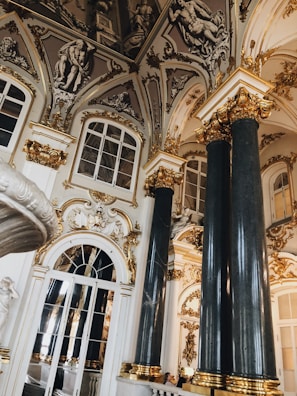 Ornate interior featuring large marble columns with gold accents and intricate moldings. The ceiling displays detailed sculptures and art, while large windows and mirrored surfaces enhance the space's grandeur. The overall design is highly decorative and opulent.