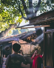 A person wearing a gray shirt with a camera hangs from their neck, capturing a photo of another person in a vibrant red dress standing in a shaded area. Sunlight filters through the leaves of a tree, creating beams that illuminate the scene. The background includes a structure with a makeshift roof, and a sign with a logo visible in the corner.