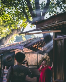 A person wearing a gray shirt with a camera hangs from their neck, capturing a photo of another person in a vibrant red dress standing in a shaded area. Sunlight filters through the leaves of a tree, creating beams that illuminate the scene. The background includes a structure with a makeshift roof, and a sign with a logo visible in the corner.