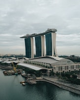 A futuristic, iconic building with three tall towers supporting a boat-shaped structure at the top, set against a cloudy sky. The waterfront area features a curved promenade and construction activity, indicating urban life and development.
