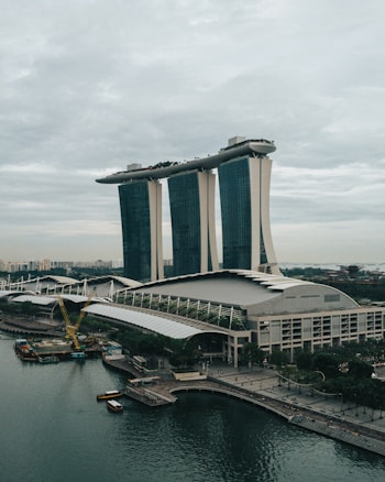A futuristic, iconic building with three tall towers supporting a boat-shaped structure at the top, set against a cloudy sky. The waterfront area features a curved promenade and construction activity, indicating urban life and development.