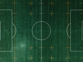 An aerial view of a soccer field with clearly defined white boundary lines and center circle on deep green artificial turf. Additional orange markings intersect the field at various points.