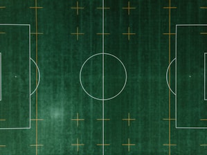 An aerial view of a soccer field with clearly defined white boundary lines and center circle on deep green artificial turf. Additional orange markings intersect the field at various points.