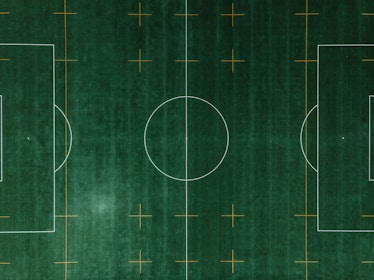 An aerial view of a soccer field with clearly defined white boundary lines and center circle on deep green artificial turf. Additional orange markings intersect the field at various points.