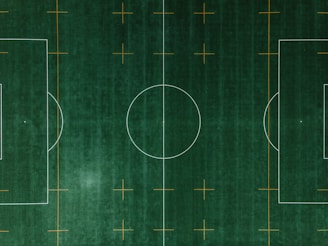 An aerial view of a soccer field with clearly defined white boundary lines and center circle on deep green artificial turf. Additional orange markings intersect the field at various points.