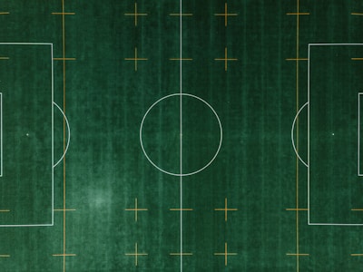 An aerial view of a soccer field with clearly defined white boundary lines and center circle on deep green artificial turf. Additional orange markings intersect the field at various points.