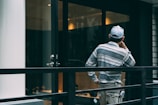 A person wearing a striped shirt and cap is standing on a balcony or terrace, facing away and appearing deep in thought. The setting includes a glass door, metal railing, and reflective surfaces suggestive of an urban or modern environment.