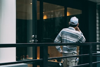 A person wearing a striped shirt and cap is standing on a balcony or terrace, facing away and appearing deep in thought. The setting includes a glass door, metal railing, and reflective surfaces suggestive of an urban or modern environment.
