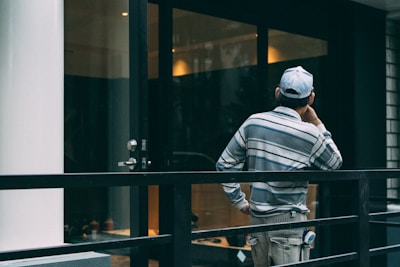 A person wearing a striped shirt and cap is standing on a balcony or terrace, facing away and appearing deep in thought. The setting includes a glass door, metal railing, and reflective surfaces suggestive of an urban or modern environment.