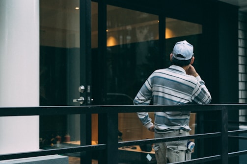A person wearing a striped shirt and cap is standing on a balcony or terrace, facing away and appearing deep in thought. The setting includes a glass door, metal railing, and reflective surfaces suggestive of an urban or modern environment.