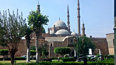 A large mosque with multiple minarets and domes, surrounded by trees and greenery in a park-like setting. There are some individuals walking and a few vehicles are seen near the structure.
