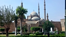 A large mosque with multiple minarets and domes, surrounded by trees and greenery in a park-like setting. There are some individuals walking and a few vehicles are seen near the structure.