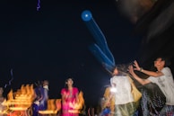 A group of people appears to be playing with a large, inflatable blue object during nighttime. The atmosphere is lively, with motion blur effects and streaks of colored lights, suggesting movement and excitement.