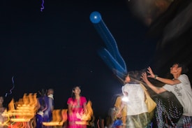 A group of people appears to be playing with a large, inflatable blue object during nighttime. The atmosphere is lively, with motion blur effects and streaks of colored lights, suggesting movement and excitement.