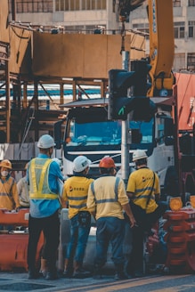 A group of construction workers wearing safety helmets and high-visibility vests are gathered around a worksite. They appear to be engaged in a discussion or task near a large truck and construction equipment. The background shows partially constructed buildings and various construction materials.