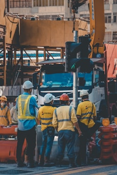 A group of construction workers wearing safety helmets and high-visibility vests are gathered around a worksite. They appear to be engaged in a discussion or task near a large truck and construction equipment. The background shows partially constructed buildings and various construction materials.