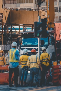A group of construction workers wearing safety helmets and high-visibility vests are gathered around a worksite. They appear to be engaged in a discussion or task near a large truck and construction equipment. The background shows partially constructed buildings and various construction materials.