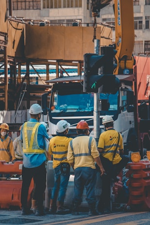 A group of construction workers wearing safety helmets and high-visibility vests are gathered around a worksite. They appear to be engaged in a discussion or task near a large truck and construction equipment. The background shows partially constructed buildings and various construction materials.