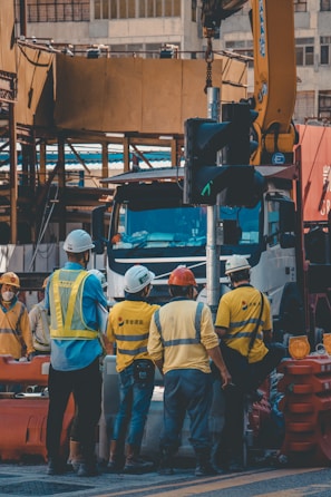 A group of construction workers wearing safety helmets and high-visibility vests are gathered around a worksite. They appear to be engaged in a discussion or task near a large truck and construction equipment. The background shows partially constructed buildings and various construction materials.