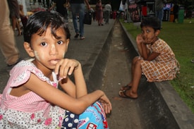 Two young children are sitting across from each other on the edge of a concrete drainage channel. The child in the foreground has traditional face paint and wears a patterned dress with polka dots and floral designs. The child in the background, wearing a brown checkered dress, rests their chin on their hands, sitting on the curb. People are walking in the background near stalls and greenery.