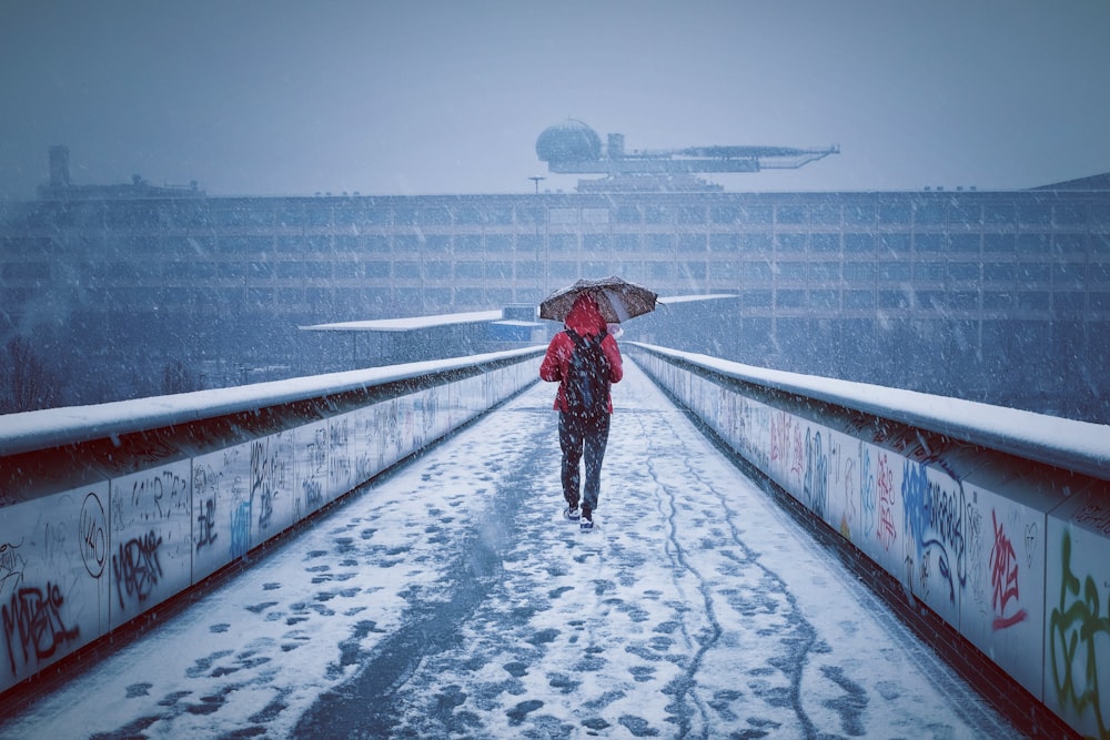 Homem com capuz vermelho e guarda-chuva caminha em caminho coberto de neve