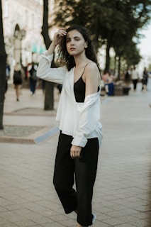 A person with curly hair stands confidently in an urban setting, wearing a white shirt over a black top and black pants. The background features a city street with trees and blurred figures in motion, suggesting a lively atmosphere.