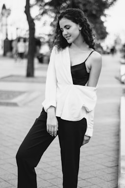 A young woman stands casually on a paved pathway in an urban setting, with her curly hair and relaxed posture conveying a sense of ease and confidence. She wears a white, off-shoulder top draped over a black top and black pants. The background is blurred, featuring out-of-focus trees and passersby.