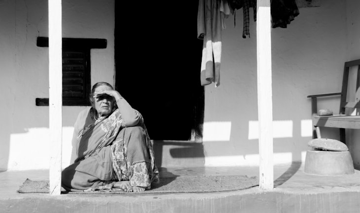 An elderly black woman with long black hair braided with roses, sitting peacefully in a rocking chair on a porch at dusk.