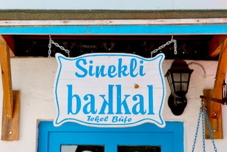 A wooden sign with blue lettering hangs above a blue door. The sign reads 'Sinekli bakkal Tekel Büfe' and is attached to chains. There is a vintage-style wall lantern on the right side, mounted on a white wall.