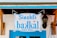 A wooden sign with blue lettering hangs above a blue door. The sign reads 'Sinekli bakkal Tekel Büfe' and is attached to chains. There is a vintage-style wall lantern on the right side, mounted on a white wall.