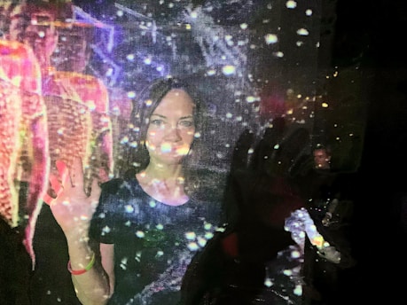 A person is standing with a hand raised, seemingly interacting with or immersed in a projection of colorful lights and patterns. The scene has a somewhat ethereal quality, with reflections and light spots scattered throughout.