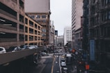 A bustling city street scene captured from an elevated vantage point, surrounded by tall urban buildings. The street is busy with traffic, including cars and buses, and there are several visible store signs and advertisements, depicting a vibrant city life.
