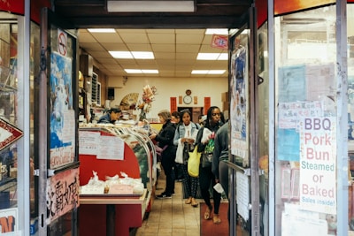 A small, bustling shop with several customers inside, including a person behind a counter attending to them. The shop is filled with a variety of items on display, such as BBQ pork buns and other food products. The entrance is adorned with signs and posters displaying information in both English and what appears to be Chinese characters. The interior has a warm and cozy atmosphere with decorations like flowers and traditional Chinese elements.