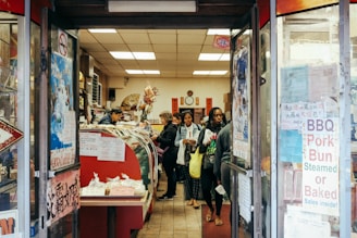 A small, bustling shop with several customers inside, including a person behind a counter attending to them. The shop is filled with a variety of items on display, such as BBQ pork buns and other food products. The entrance is adorned with signs and posters displaying information in both English and what appears to be Chinese characters. The interior has a warm and cozy atmosphere with decorations like flowers and traditional Chinese elements.