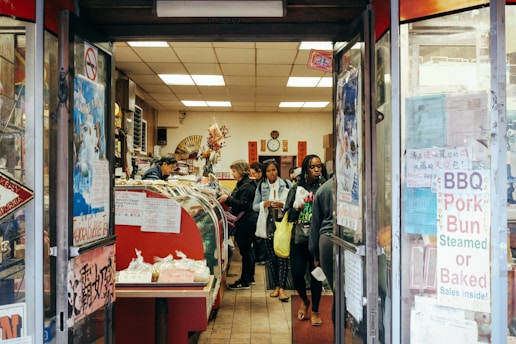 A small, bustling shop with several customers inside, including a person behind a counter attending to them. The shop is filled with a variety of items on display, such as BBQ pork buns and other food products. The entrance is adorned with signs and posters displaying information in both English and what appears to be Chinese characters. The interior has a warm and cozy atmosphere with decorations like flowers and traditional Chinese elements.