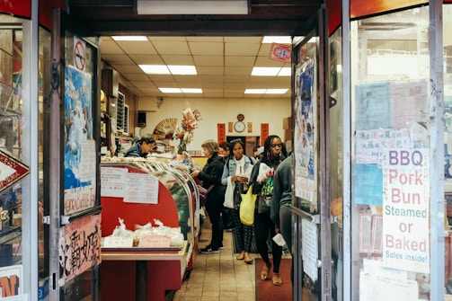 A small, bustling shop with several customers inside, including a person behind a counter attending to them. The shop is filled with a variety of items on display, such as BBQ pork buns and other food products. The entrance is adorned with signs and posters displaying information in both English and what appears to be Chinese characters. The interior has a warm and cozy atmosphere with decorations like flowers and traditional Chinese elements.