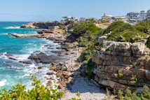A coastal landscape featuring rugged cliffs and rocky shorelines with turquoise blue ocean waves crashing against them. Along the top of the cliffs, there are green shrubs and trees, with a few buildings and a parking area visible in the background. People can be seen walking along paths and standing on the rocks, enjoying the scenic view.