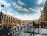 A bustling indoor courtyard resembling an Italian architecture with ornate buildings and arched windows. People are walking around and ascending a grand staircase. Elegant street lamps are lit, adding to the ambiance. The ceiling is painted to mimic a blue sky with soft, fluffy clouds.