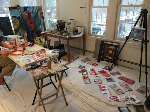 A bright, creative studio space featuring a table covered with various art supplies, including paints, glue, and paper cutouts. On the floor, a white sheet displays an array of colorful handmade cards and artwork. An easel holds a canvas with an abstract painting, and the walls are lined with large paintings, some leaning on the floor. The room is well-lit by three large windows in the background, allowing natural light to fill the space.