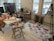 A bright, creative studio space featuring a table covered with various art supplies, including paints, glue, and paper cutouts. On the floor, a white sheet displays an array of colorful handmade cards and artwork. An easel holds a canvas with an abstract painting, and the walls are lined with large paintings, some leaning on the floor. The room is well-lit by three large windows in the background, allowing natural light to fill the space.