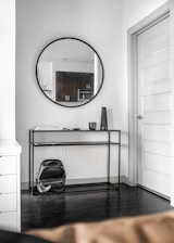 The image features a modern, minimalist interior corner. A round mirror hangs on a white wall above a sleek, black metal console table. On the console are a few items, including a small cylindrical object and some papers, adding to the minimalist aesthetic. A black backpack is placed on the floor underneath the table. The room has a simple, clean design with a closed white door on the right and dark hardwood flooring.
