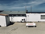 A beachside structure with a white exterior stands on a sandy area. A propane tank labeled 'WESTGAS IEPER' is positioned in front of the building. The roof is metal, and a clear blue sky and distant ocean are visible in the background.