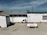A beachside structure with a white exterior stands on a sandy area. A propane tank labeled 'WESTGAS IEPER' is positioned in front of the building. The roof is metal, and a clear blue sky and distant ocean are visible in the background.