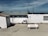A beachside structure with a white exterior stands on a sandy area. A propane tank labeled 'WESTGAS IEPER' is positioned in front of the building. The roof is metal, and a clear blue sky and distant ocean are visible in the background.