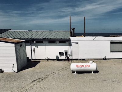 A beachside structure with a white exterior stands on a sandy area. A propane tank labeled 'WESTGAS IEPER' is positioned in front of the building. The roof is metal, and a clear blue sky and distant ocean are visible in the background.